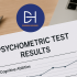 Hand holding a psychometric test results report with charts on cognitive abilities, personality traits, and job fit, placed on a wooden desk next to a laptop.